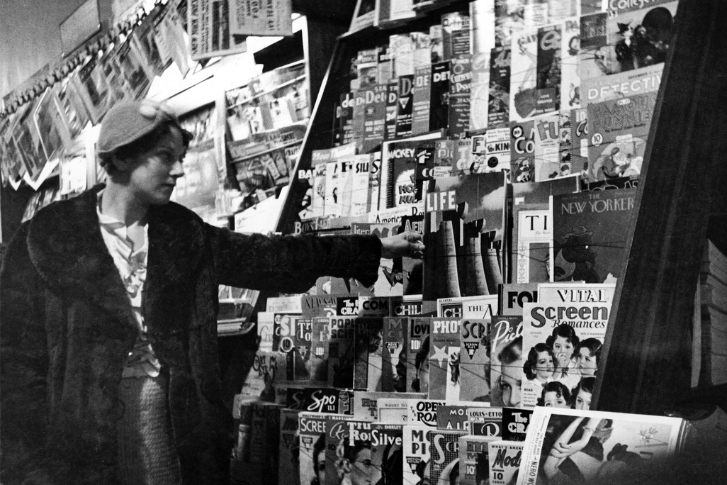 A black and white image of a young woman, wearing a cap and fur coat, reaching for a copy of Life on a crowded newsstand; a copy of the New Yorker and Time are also in view