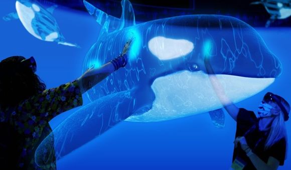 Two people an augmented-reality experience wear headsets while reaching out to "touch" a holographic killer whale.