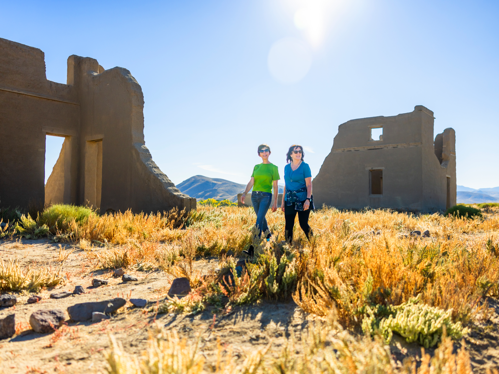 Two people walk through a field of dry grass and shrubs toward the ruins of old, crumbling buildings under a clear, sunny sky, with distant mountains in the background.