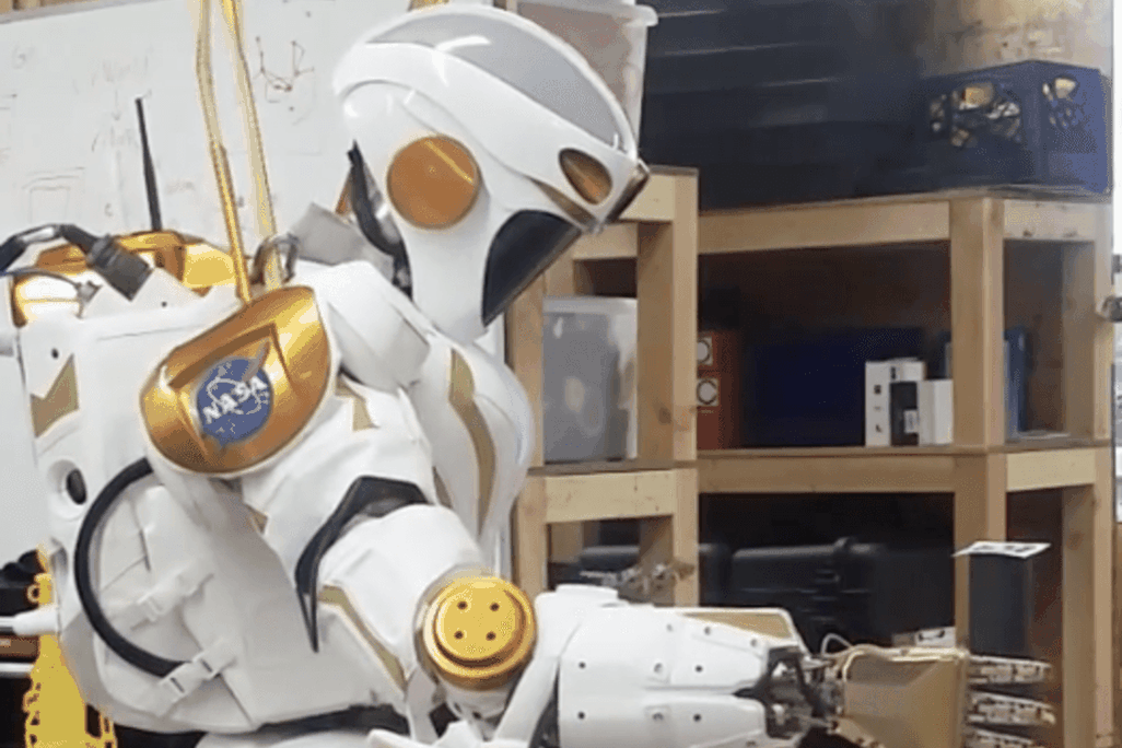 A NASA Valkyrie robot picks up an item with its hand.