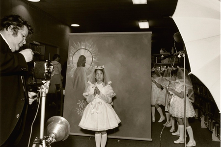 Black and white photograph of a girls posing for a first communion picture. A dropdown background image of a man with a halo around his head. A photographer stands with a camera in front of the girl.