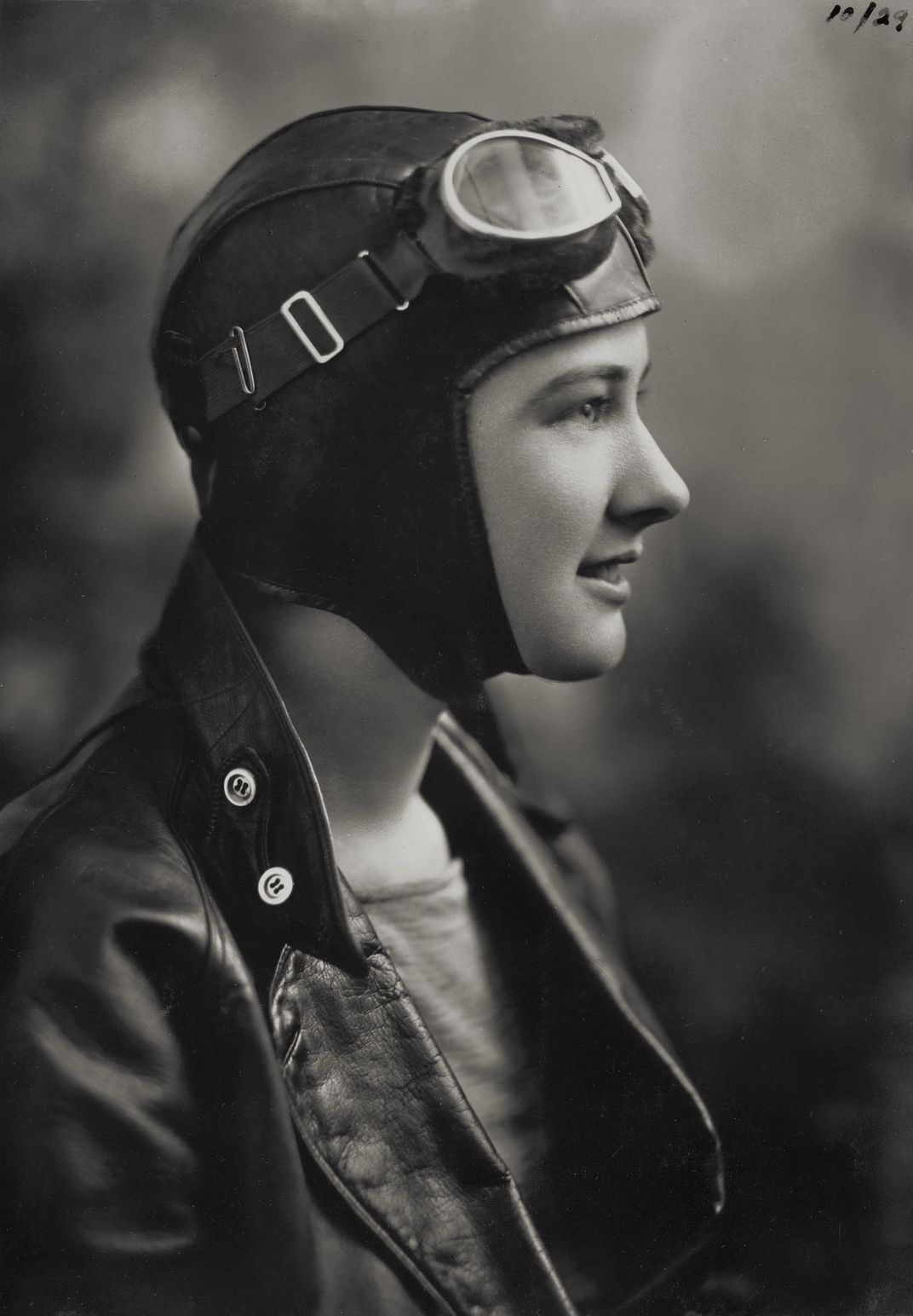 Profile of a woman wearing a flying cap with goggles resting on top. She wears a leather collared jacket