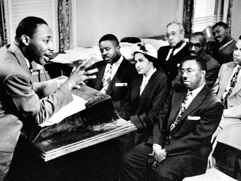 MLK Jr stands at a podium and speaks animatedly to a group of seven other people in chairs, including Rosa Parks and Ralph Abernathy, who all sit facing him