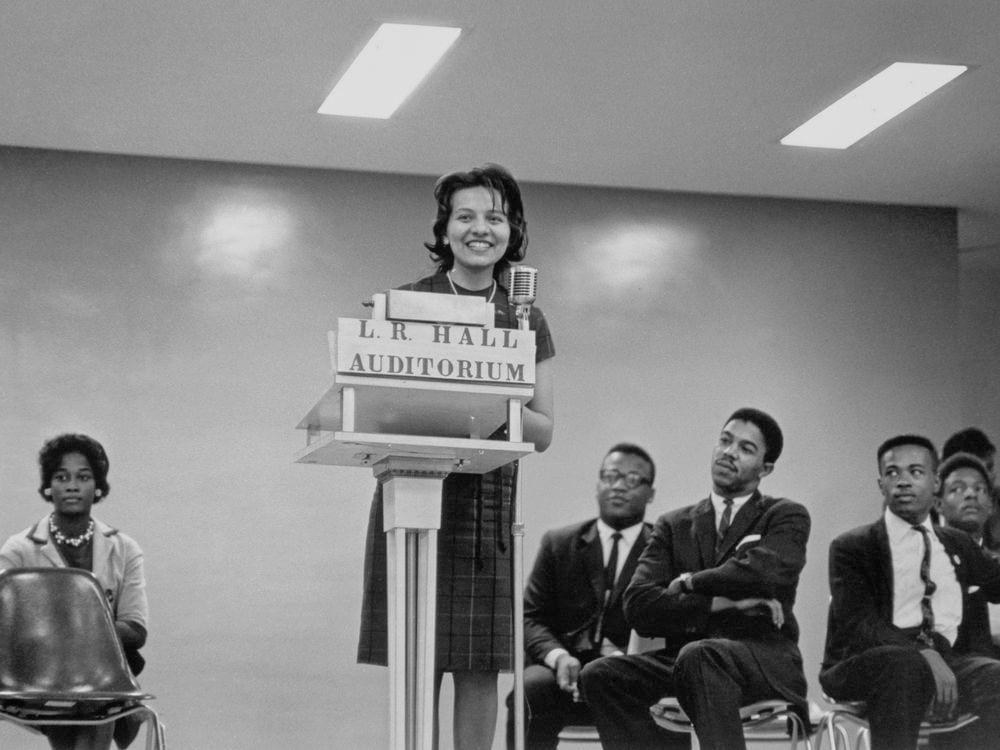 A black-and-white image of Diane Nash at the podium with men and a woman sitting on either side of her in chairs. Nash stands in the center of the image behind the podium labeled [L.R. HALL/ AUDITORIUM] speaking into the microphone.