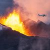 Red hot molten lava and thick dark dust spew from the ground while a small white. single-propeller aircraft flies overhead.