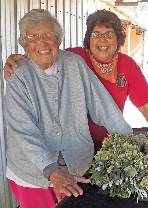 Two women who look very similar, except one is elderly, both with short wavy hair and glasses, smile and pose next to an elaborate green floral arrangement.