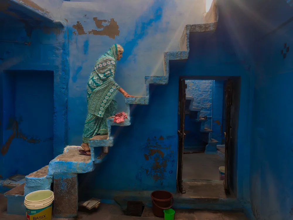 An elderly woman, dressed in a patterned green and white sari, ascends a rugged, blue-painted staircase in what appears to be a courtyard or interior space in Jodhpur, India. The walls and steps are all painted in a deep, vibrant blue, characteristic of the city's old quarter. Sunlight streams down from above and to the right, illuminating the woman and casting dramatic highlights and shadows on the textured walls. A doorway leads to another blue-walled area below, and various household buckets and containers are visible near the bottom of the stairs.