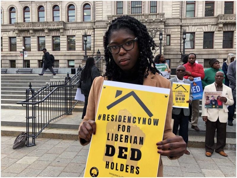 Woman stands in front of a group of protestors outside of a courthouse. She is holding a yellow protest sign that reads, "#ResidencyNow for Liberian DED Holders."