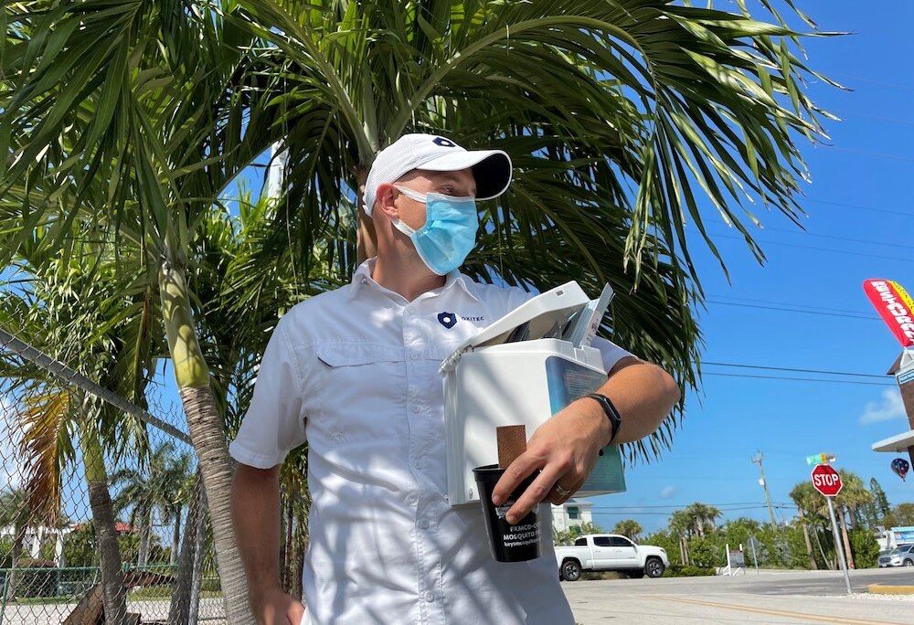 A man wearing a blue mask and a white baseball cap stands in front of a palm tree and holds a hexagon-shaped, toaster-sized box and a black cup