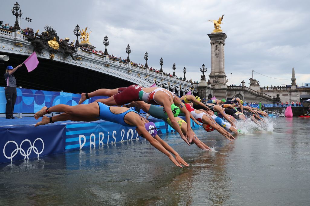 A line of athletes diving into a river with a bridge in the background