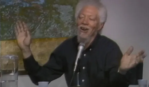 Benny Andrews sits at a table with a microphone, cups, and a pitcher of water with a yellow, green, and blue abstract painting hanging behind him. He is wearing a black button down shirt and raising his hands while talking.