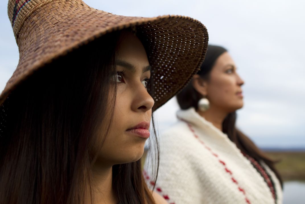 A close-up of a woman in a hat. Another woman in a white sweater stands behind her. 