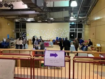 A large, multipurpose room with a variety of tables that display student work. Adults stand at each table. A sign in the foreground reads, “This way to the exhibits”.