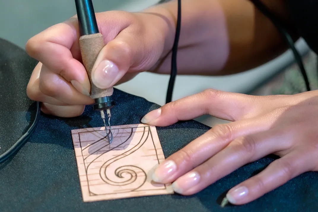 Close-up on two hands working with a pen-like wood-burning tool, etching dark curving lines into a small rectangular piece of tan park. The image resembles a wave.