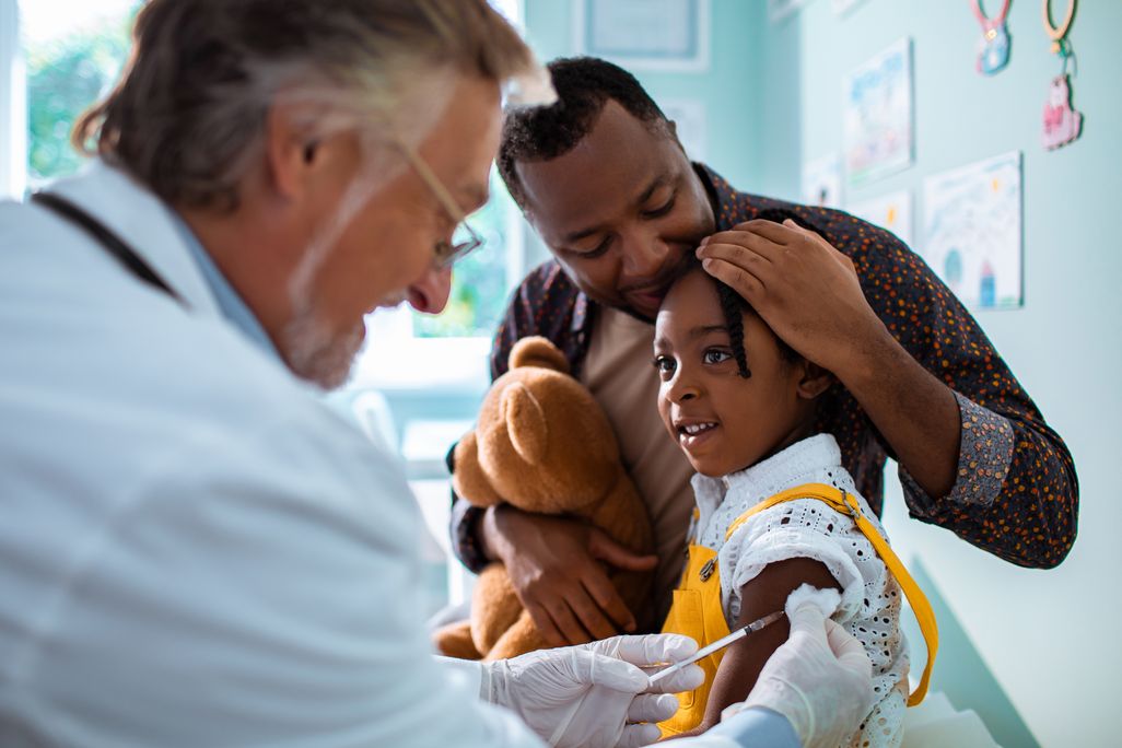 An image of a older male physican vaccinating a young child. The child is smiling and is being held by her father.