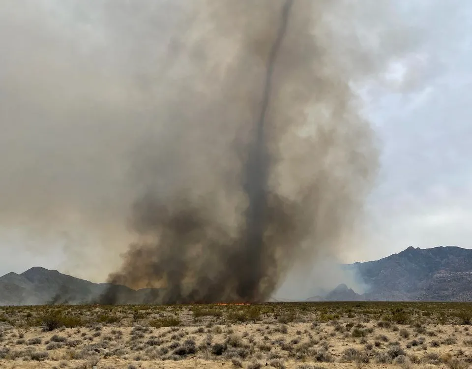 a column of black smoke rises in a desert with mountains in the background