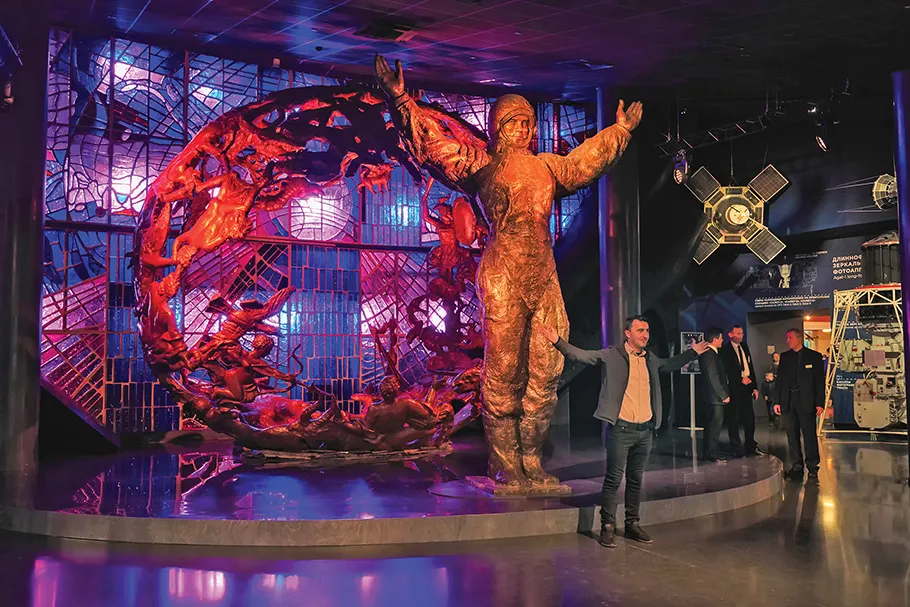 A museum display features a statue of a male astronaut standing with his arms outstretched in front of a metal sculpture lit by red and purple lighting. A museum visitor stands in a similar pose in front of the statue.