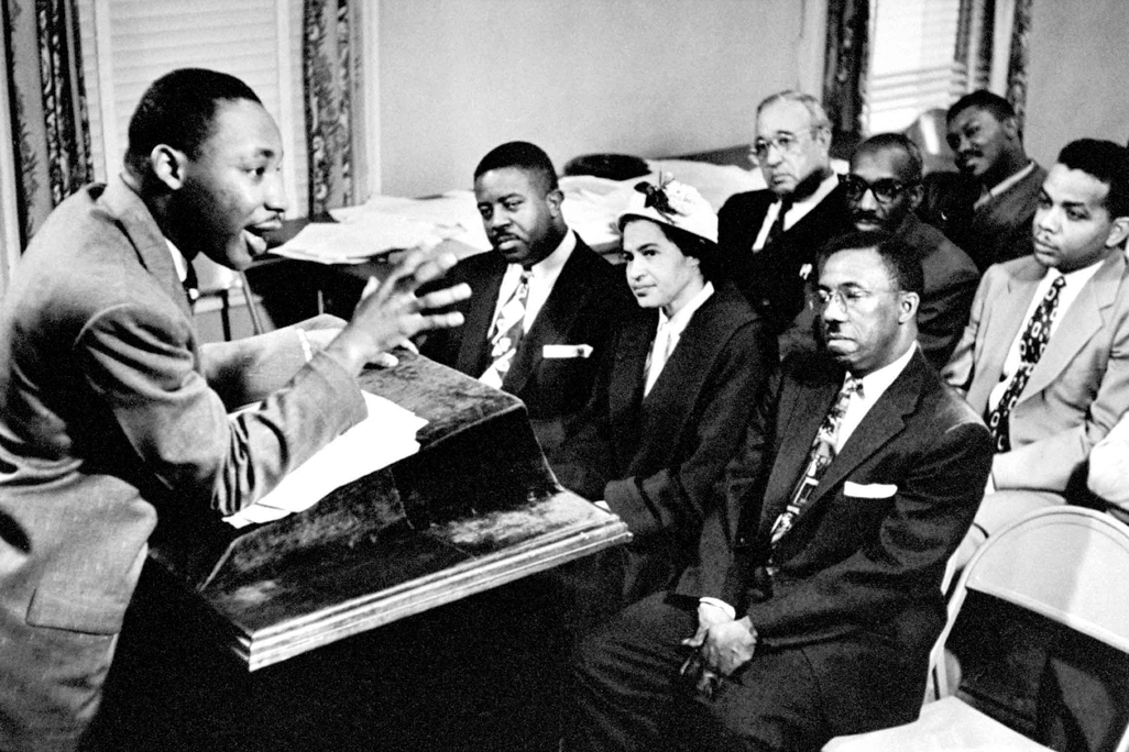 MLK Jr stands at a podium and speaks animatedly to a group of seven other people in chairs, including Rosa Parks and Ralph Abernathy, who all sit facing him