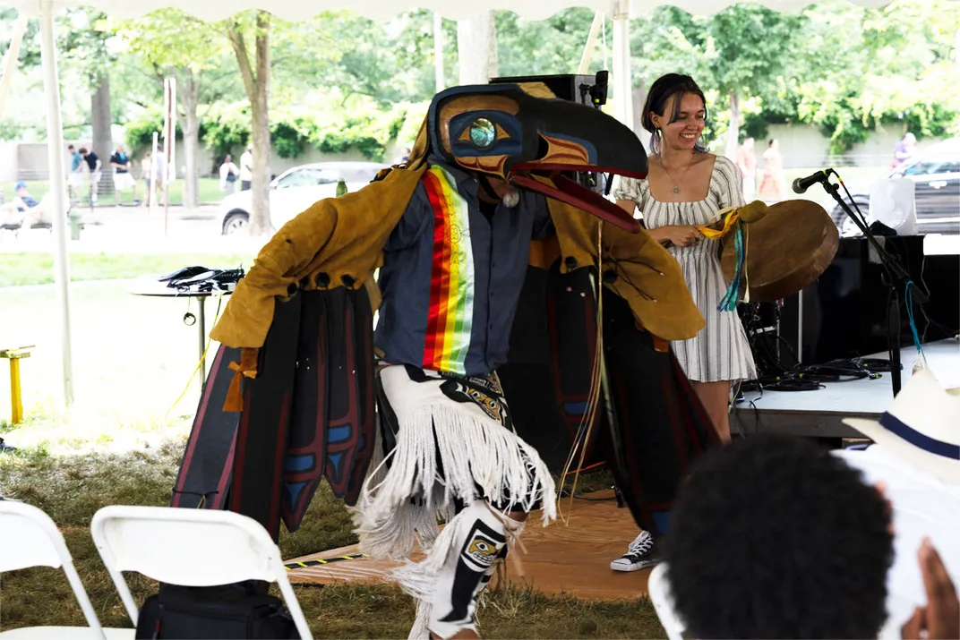 A person wearing feathered regalia and a birdlike wooden mask dances outdoors.