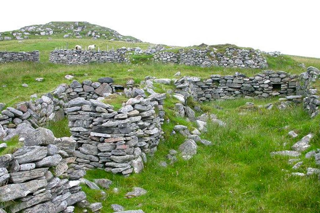 A field of green grass with a glimpse of grey sky behind; in the foreground, low stacked walls of flat rocks form the remains of terraces built into a sloping hill