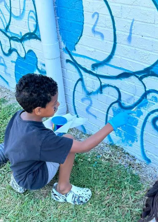 A child crouches in the grass to paint in blue shapes on a brick wall.