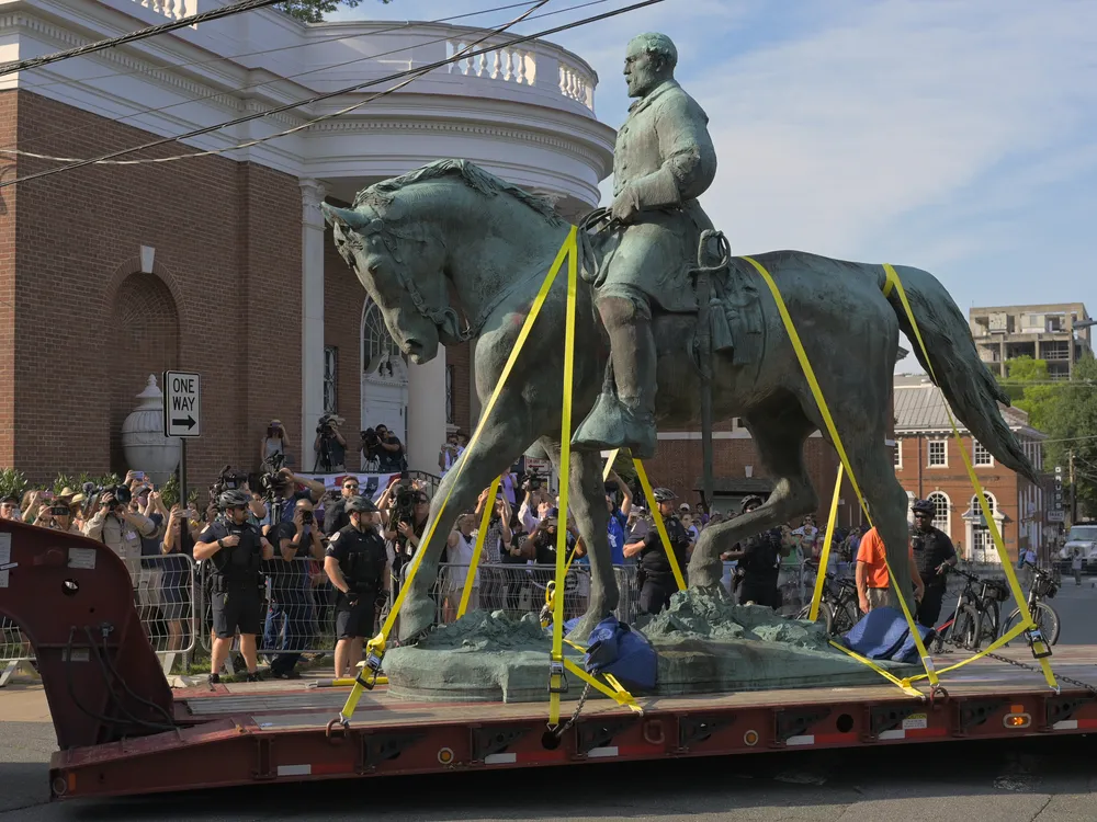 Construction crews transport a greenish bronze statue of Lee on horseback past a crowd of onlookers on a truck bed