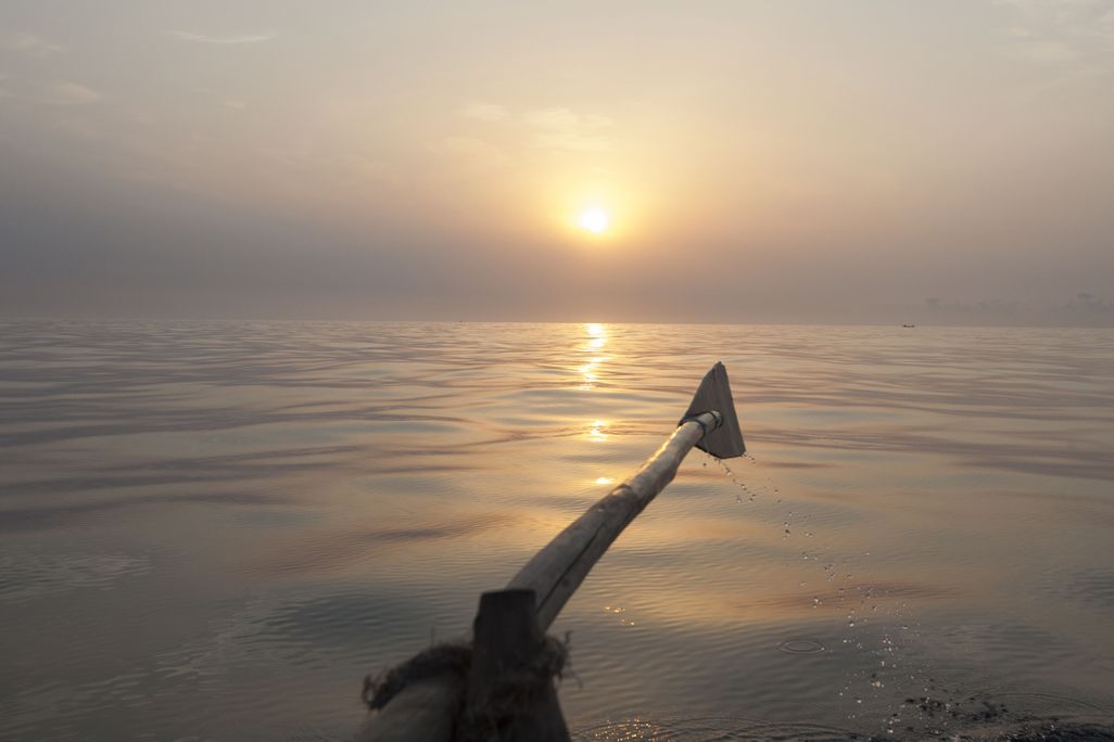A view of an oar raised parallel to the water as the sun slowly sets