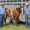 Two men in blue shirts, blue jeans, leather belts, and white cowboy hats pose in front of a brown horse.