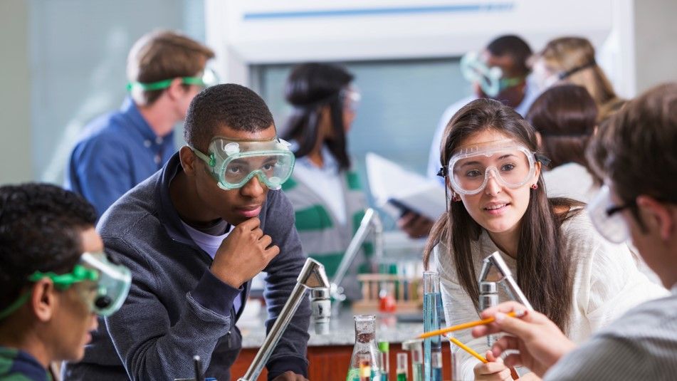 Young adult students of various ethnic backgrounds doing a chemistry experiment in class. The students are all wearing protective safety goggles.
