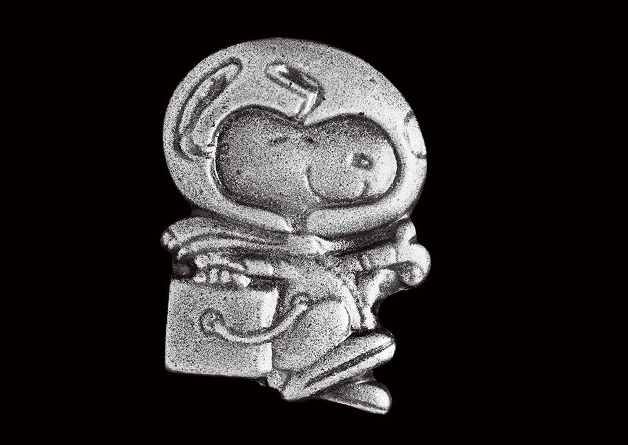 A small silver pin shows a smiling Snoopy, a cartoon dog, wearing a spacesuit with a large clear glass helmet, performing his iconic happy beagle dance.