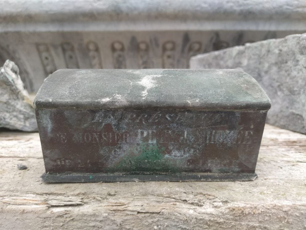 A square metal casket that is rusting and has gone green in some places, close-up, with some inscriptions that read "The heart of Pierre David was solemnly placed in the monument on 25 June 1883"