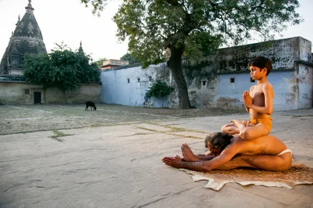 A man stretches into a west-facing extension known as paschimattanasana with his son perched on his back in padmasana, or lotus pose | Varanasi