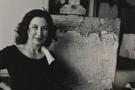 Grayscale photograph of smiling woman dressed in a black top and pants with her right hand resting on her hip and her right hand next to her neck standing in front of an abstract painting on an easel.
