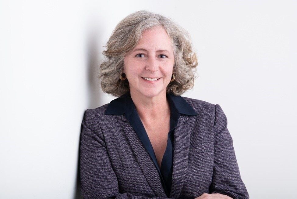 Elizabeth Babcock from the waist up against a white background. She is leaning against a wall with her arms crossed in front of herself.