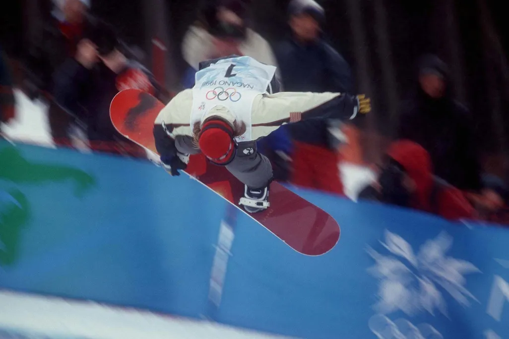 snowboarder performs trick at 1998 olympics, reaching down to touch the board