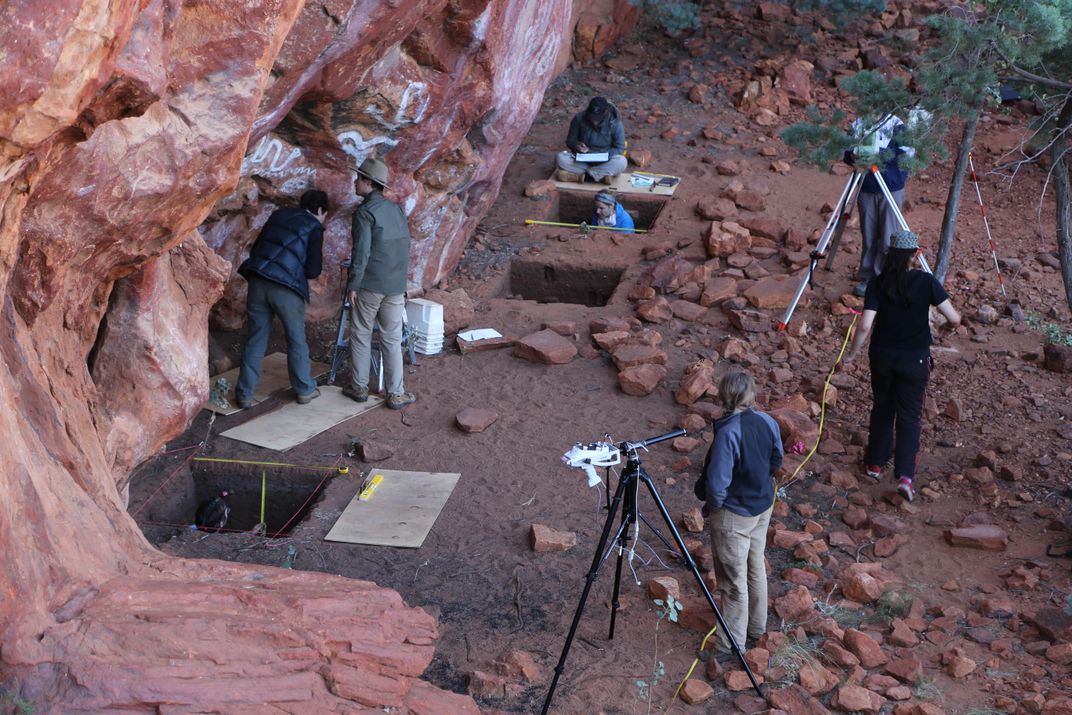 A team of archaeologists conducts field work, digging square holes and using microscopes on dark red dirt