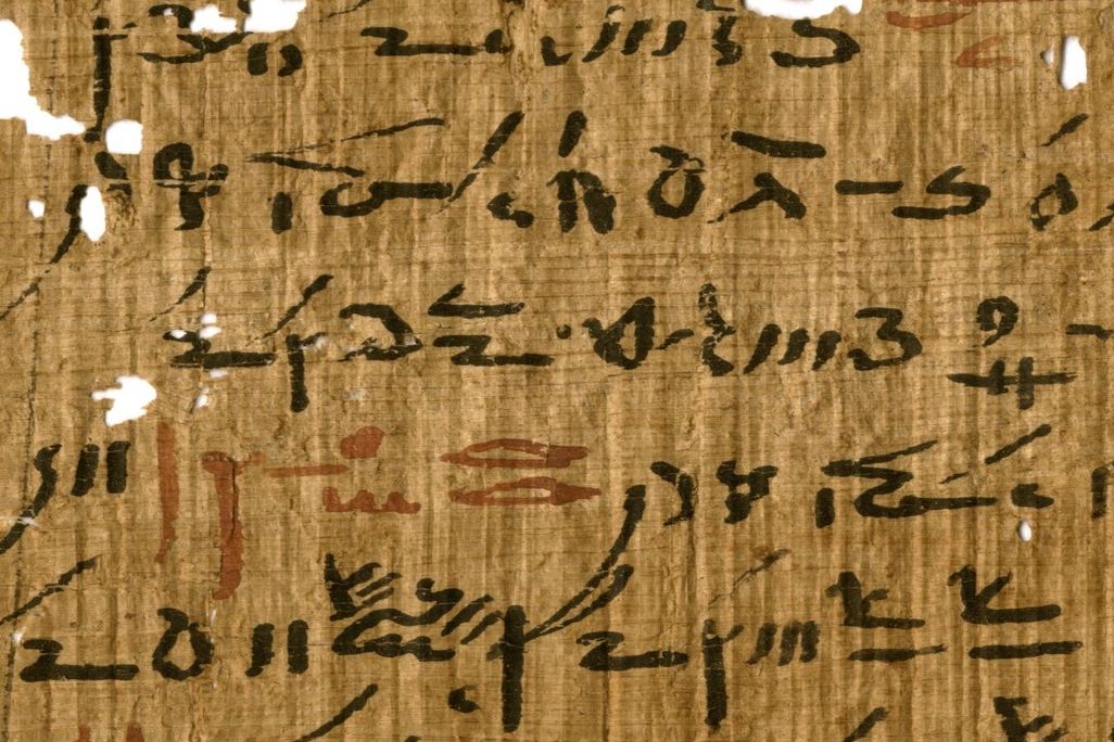 A yellow-orange papyrus sheet, with some small holes, with elongated handwritten black hieroglyphic script, with small sections every few lines written in faded red ink