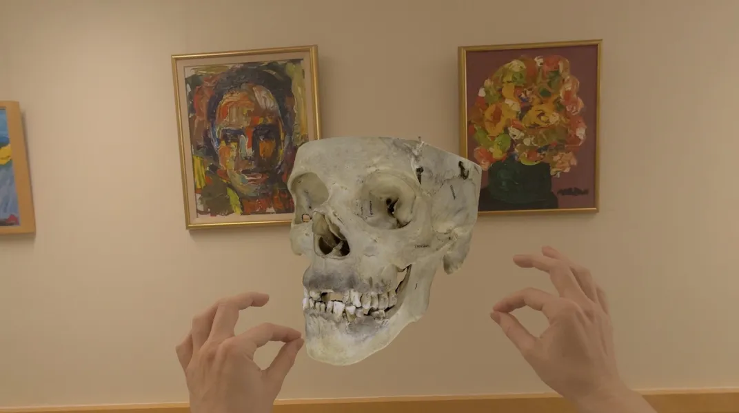 two hands pinch the air to manipulate a virtual human skull, shown floating in front of artwork on a wall