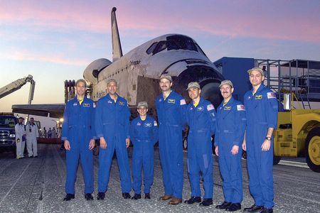 Seven people wearing blue flightsuits stand in front of a space shuttle parked outside.