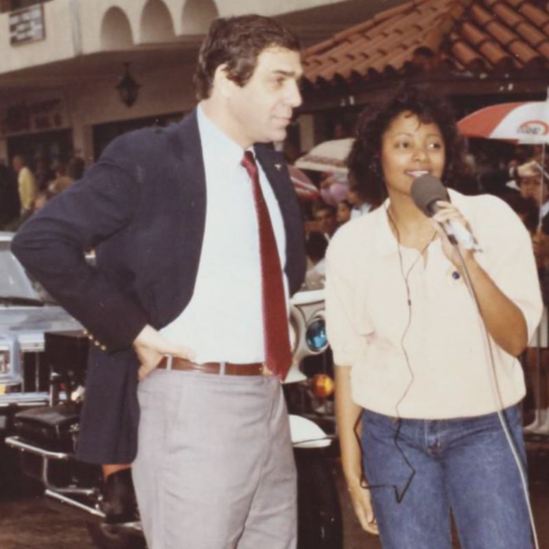 woman, Lori Montenegro, is holding a microphone and  interviewing a man. In the background are buildings and cars