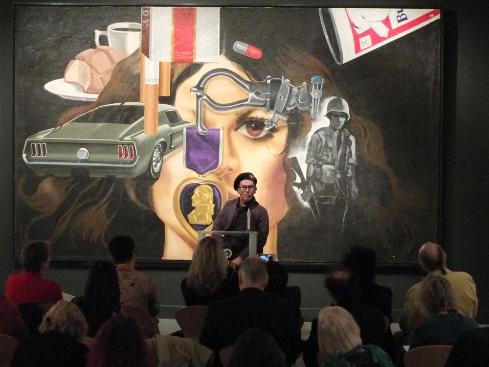 Jesse Treviño speaking in front of guests. His artwork "Mi Vida" is projected behind him.