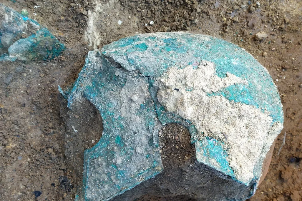 A view of a blue-green oxidized metal helmet, with sharp curved cheek plates that extend past the face, lying on the ground in the dirt