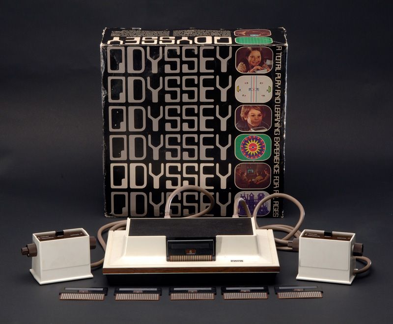 The Magnavox Odyssey system arrayed in front of its cover box. The box is decorated with the system's name and small images of various games and children using the Odyssey's controller