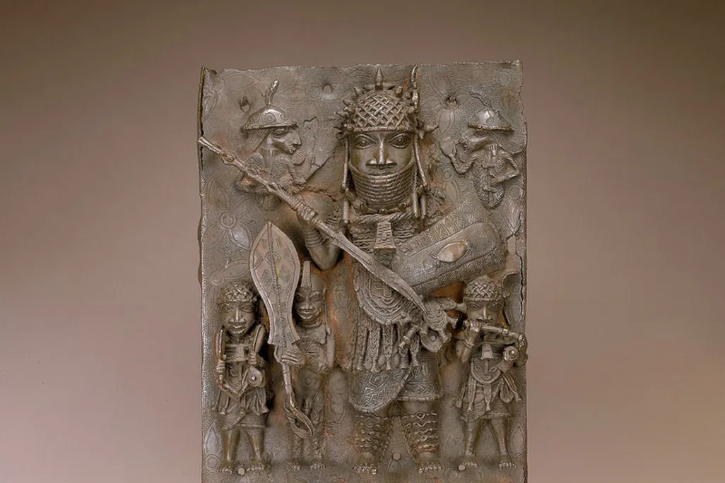 A brown metal plaque that is intricately carved with figures, including a large central figure wearing armor and towering over at least three smaller people standing beneath