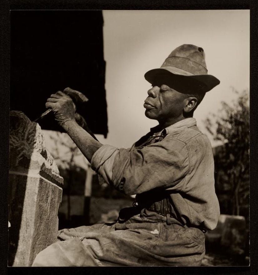 A Black man in a hat, overalls and shirt sits down and works intently on a sculpture
