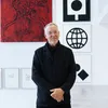 Artist Matt Mullican wearing glasses and a black jacket with his hands folded in front of his body, standing in front of six artworks, with his body partially hiding them.