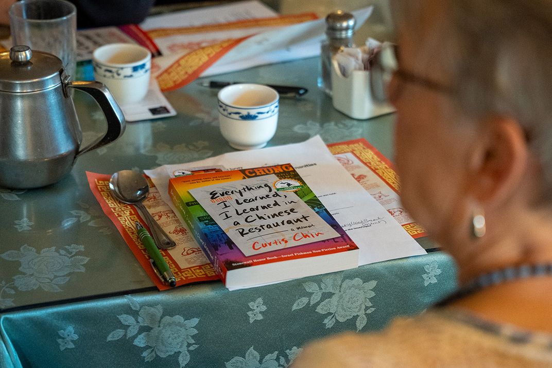 A restaurant table set with Chinese-style cups, paper placemats, seasonings, and a book: Everything I Learned, I Learned in a Chinese Restaurant by Curtis Chin. A diner sits in the foreground, out of focus.