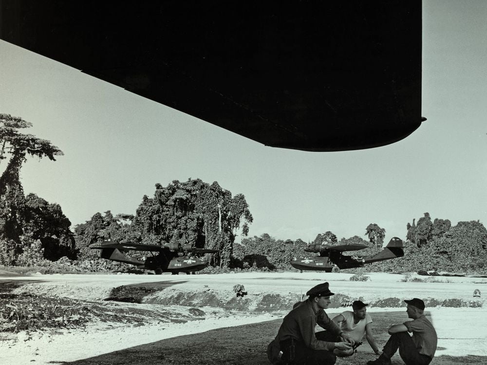 Three men sit under the shade of a massive airplane wing; one of the men is wearing an officer's hat. Two huge aircraft are parked in the background, both are painted black. Behind them is a lush island landscape. The photo has been given a green tint.