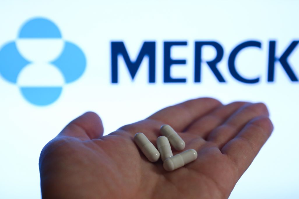 Image of left hand holding four white capsules against a Merck-branded backdrop