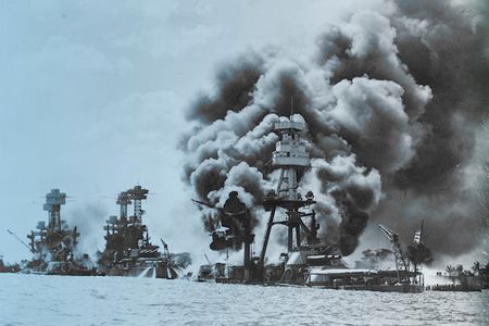 The USS Arizona Memorial floats above the ship’s sunken hull. Battleships burn in the aftermath of the attack; the Arizona is at far right.